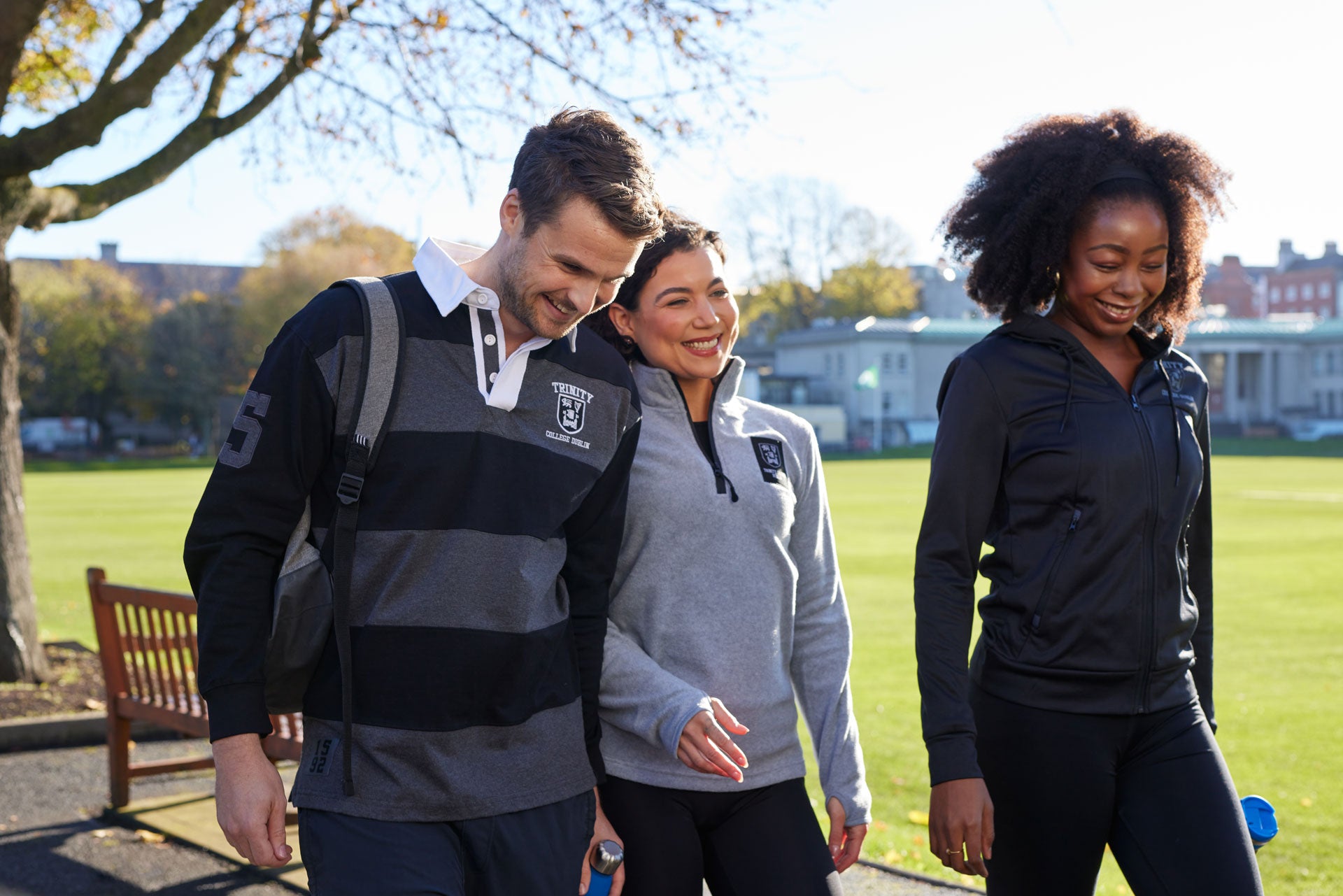 Three people walking outdoors on a sunny day, wearing Trinity College Dublin branded clothing.