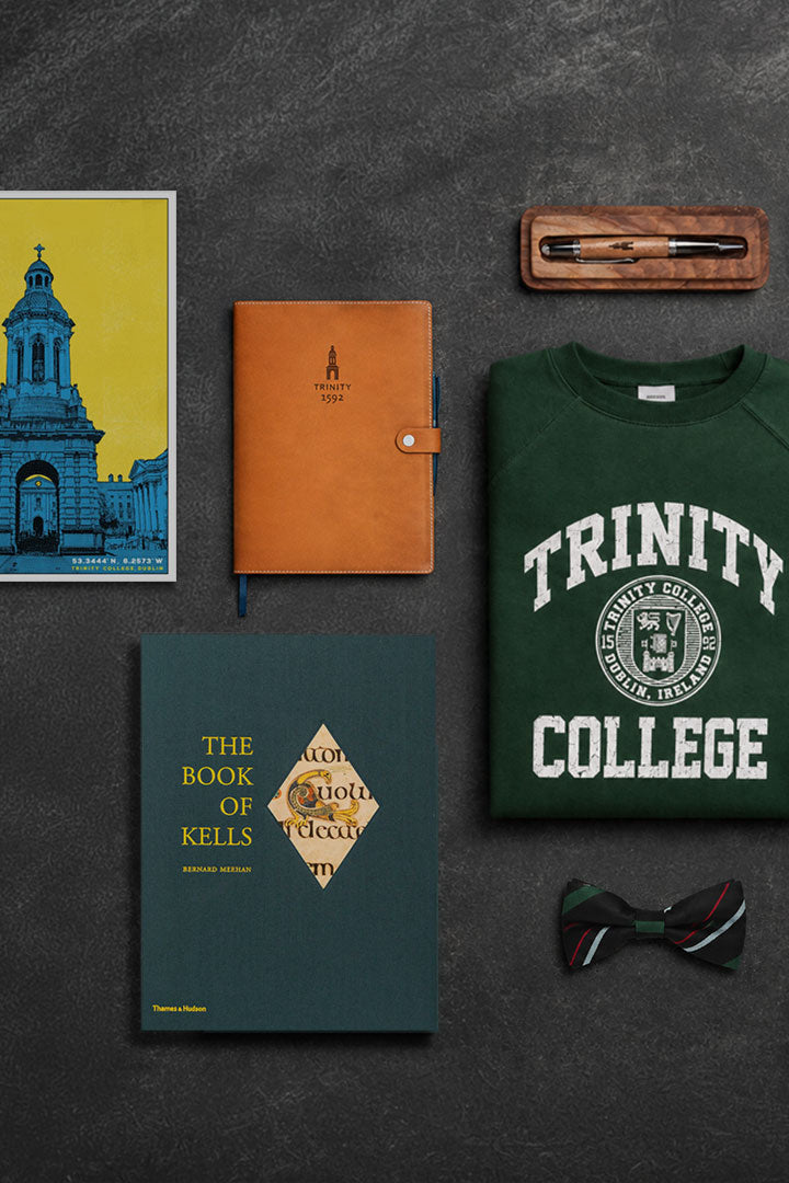 Collection of items including a green 'Trinity College' sweatshirt, a brown 1592 notebook, a blue book titled 'The Book of Kells', and a bow tie on a dark surface.