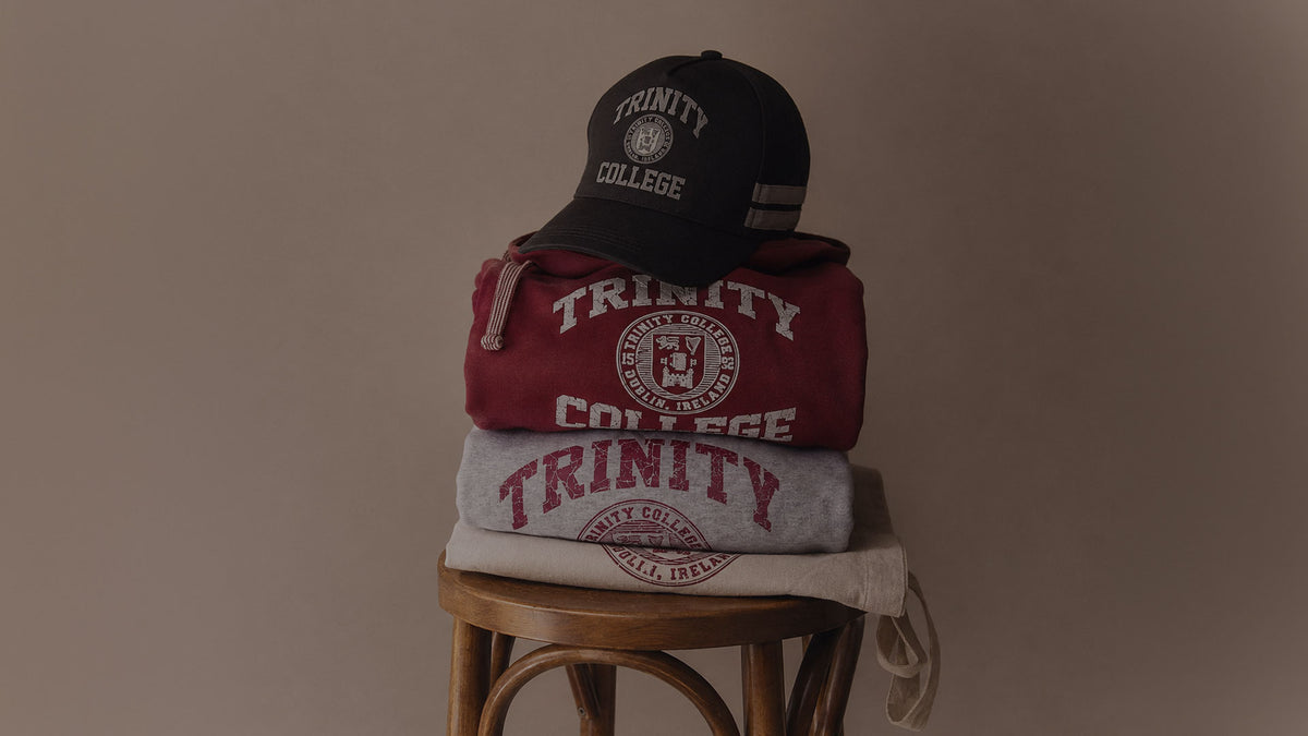 A stack of branded apparel on a wooden stool against a plain taupe wall: a black cap at the top, a dark red sweatshirt, a heather gray sweatshirt, and a cream drawstring bag at the bottom. Each item features large white 'TRINITY COLLEGE' lettering and crest graphics.