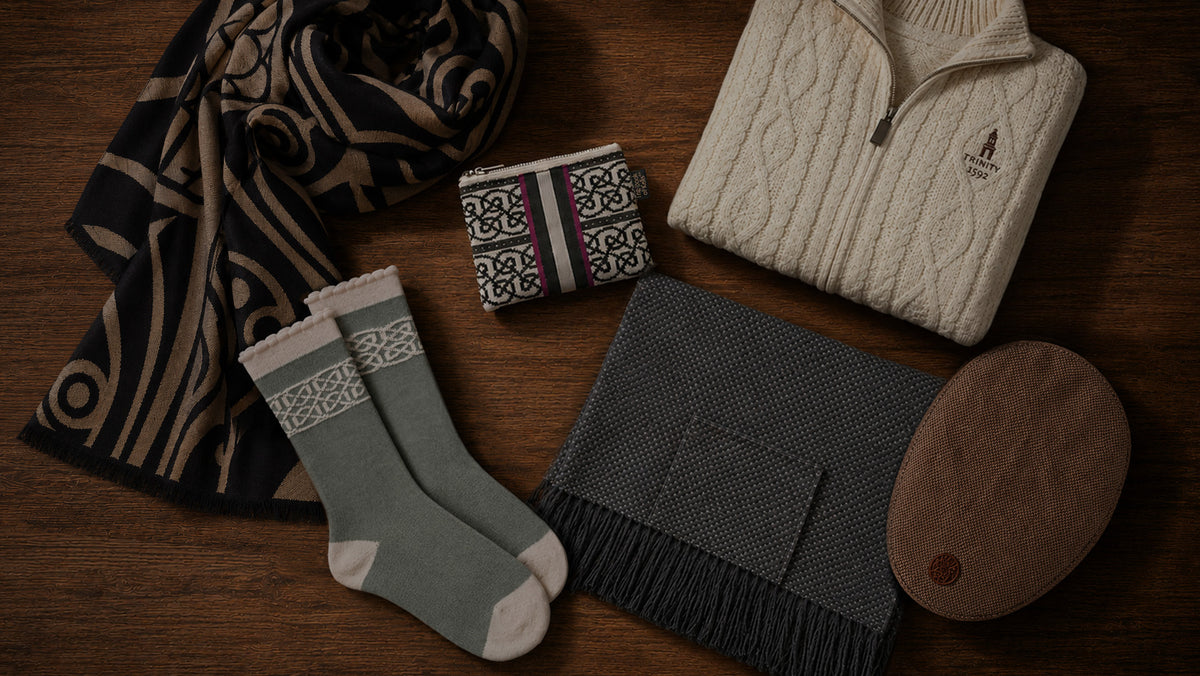 A curated flat-lay of multiple knit and textile products on a dark wood surface, including a cream cable-knit quarter-zip sweater, a dark patterned scarf, a pair of muted green crew socks with white knit cuffs, a dark gray fringed scarf, a small patterned folded item, and a brown oval leather accessory.