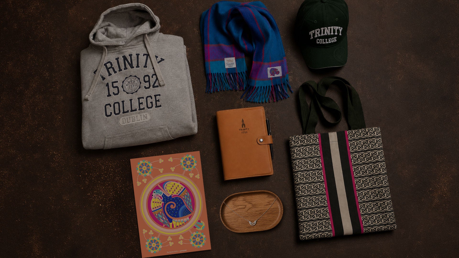 A flat-lay collection of collegiate-themed merchandise on a dark brown textured background, including a gray hoodie, a blue and purple scarf, a green baseball cap, a patterned tote bag, a leather notebook, and a colorful art print.