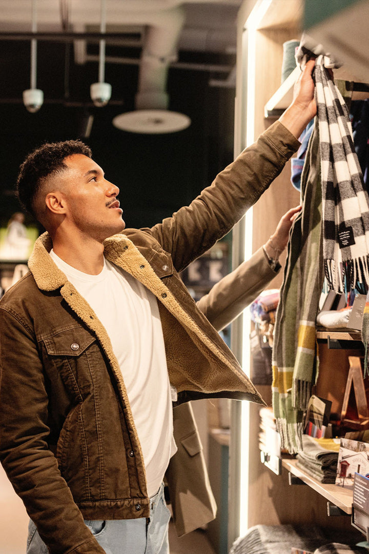 A man in a brown suede jacket with a tan shearling lining browses a clothing rack in a retail store, holding a black and white plaid scarf.