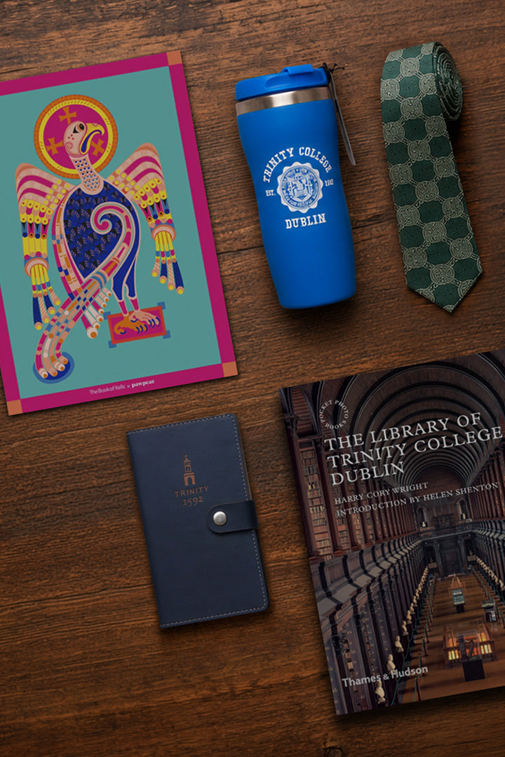 A styled flat-lay on a wooden surface featuring a colorful art print, a blue travel tumbler with Trinity College Dublin branding, a green checkered necktie, a navy leather passport cover with an embossed Trinity College logo, and a book titled 'The Library of Trinity College Dublin'.