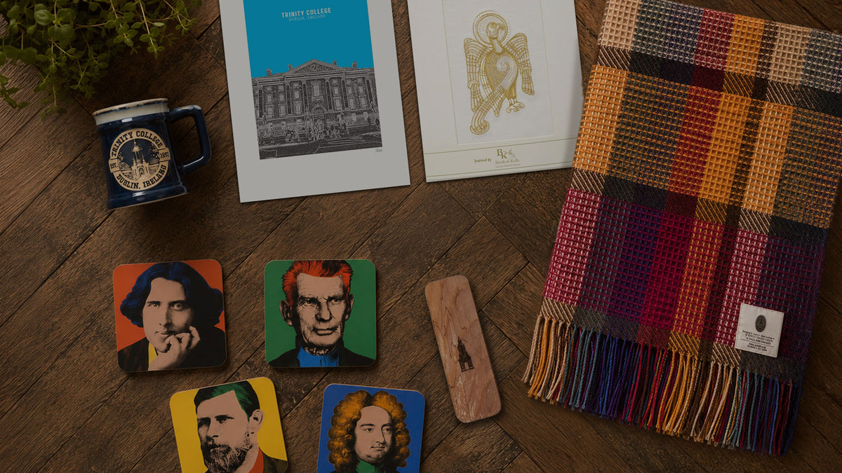A styled flat-lay of a colorful woven scarf with fringe, several square coasters featuring illustrated portraits, a small mug, printed cards, and a wooden object on a dark wood surface.