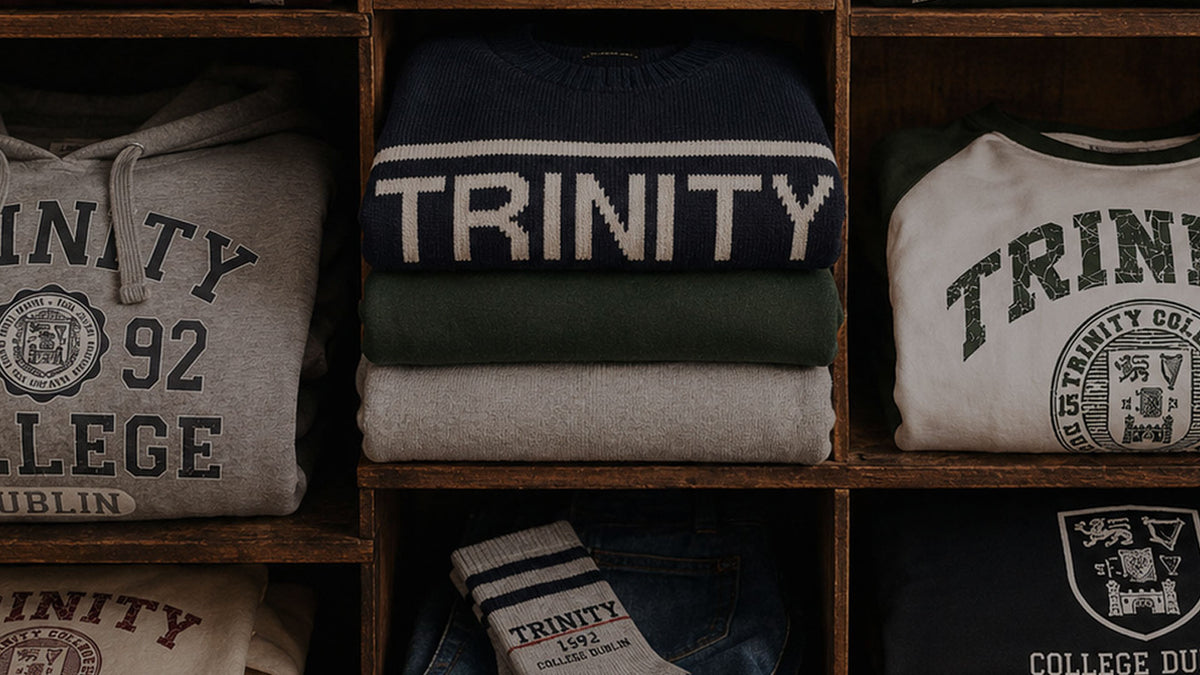 Stacked collegiate apparel folded in wooden cubby shelves, including a navy knit sweater with large white 'TRINITY' lettering, a gray hoodie with dark lettering, a cream T-shirt with dark green 'TRINITY' text, and folded jeans with a visible 'TRINITY' sock.